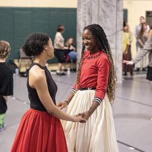 Two students in long skirts smile at each other as they hold hands.